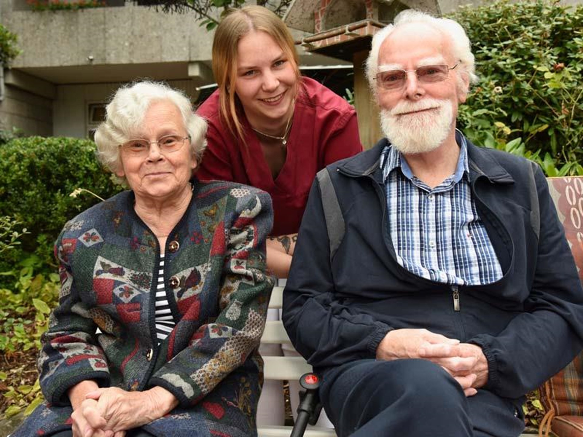 eine Seniorin und ein Senior sitzen im Garten eines Pflegeheims, eine junge Pflegerin unterstützt sie