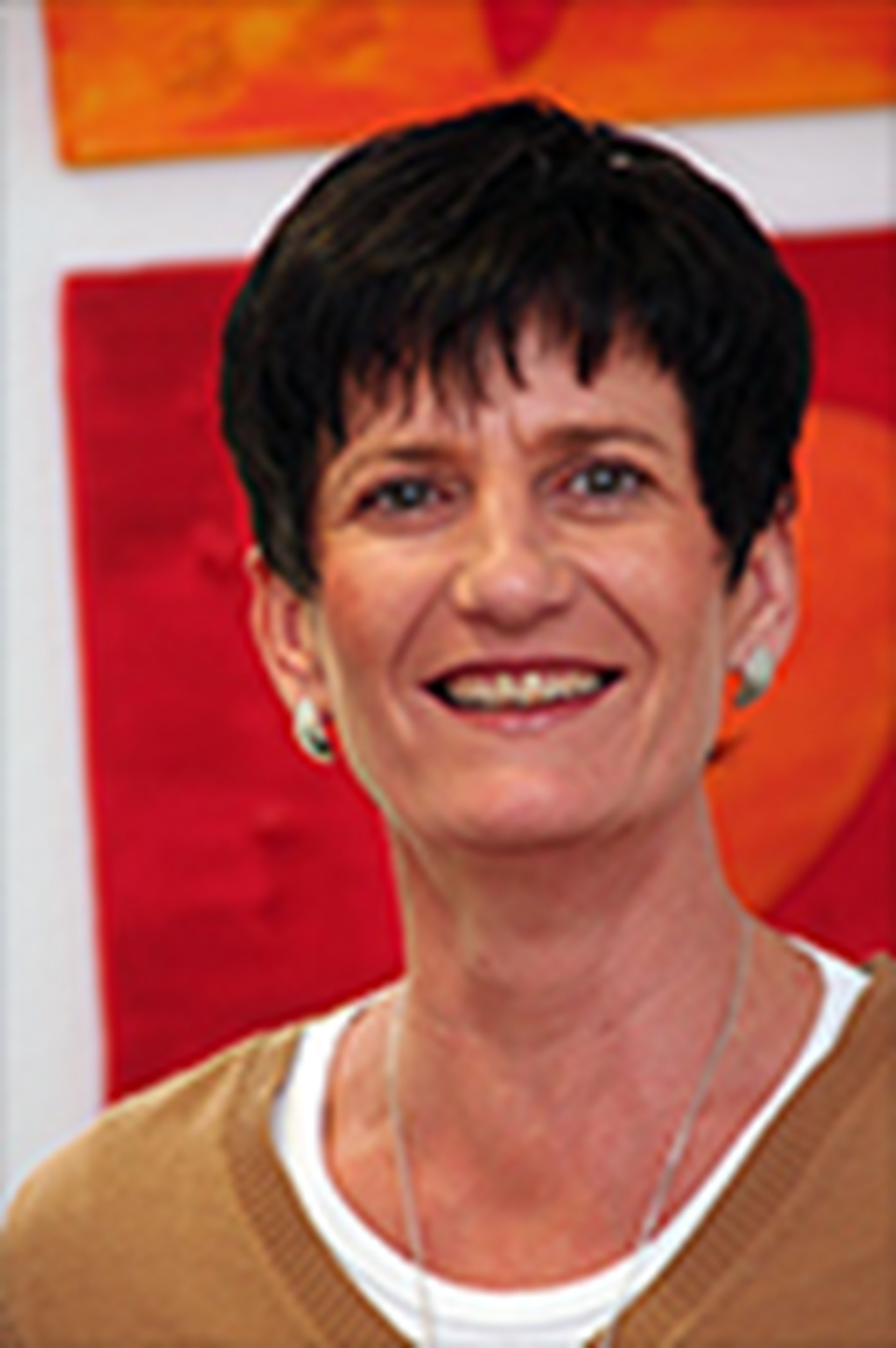 Lächelnde Frau mit schwarzen Haaren, die ein weißes T-Shirt mit einem braunen Pullover trägt und vor einem gelb-roten Gemälde steht