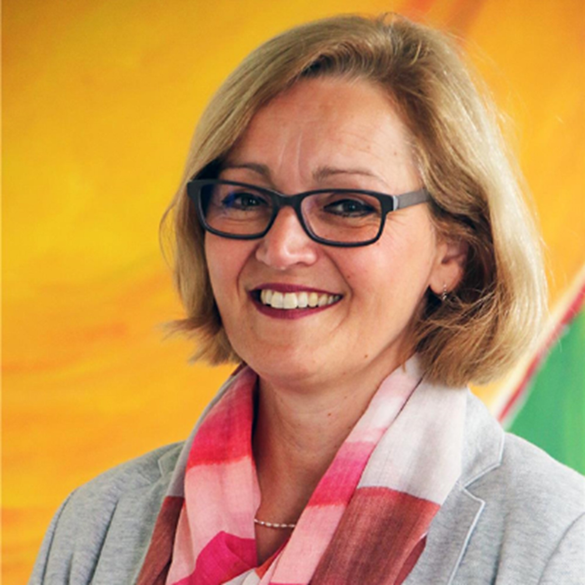 Lächelnde Frau mit schulterlangen, blonden Haaren und Brille, die einen rötlich-gestreiften Schal und eine hellgraue Anzugjacke trägt. Sie steht vor einem gelben Hintergrund.