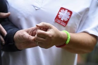 Pflegerin in weißem Shirt mit Flammenkreuz hilft alter Dame, konzentriert auf Hände