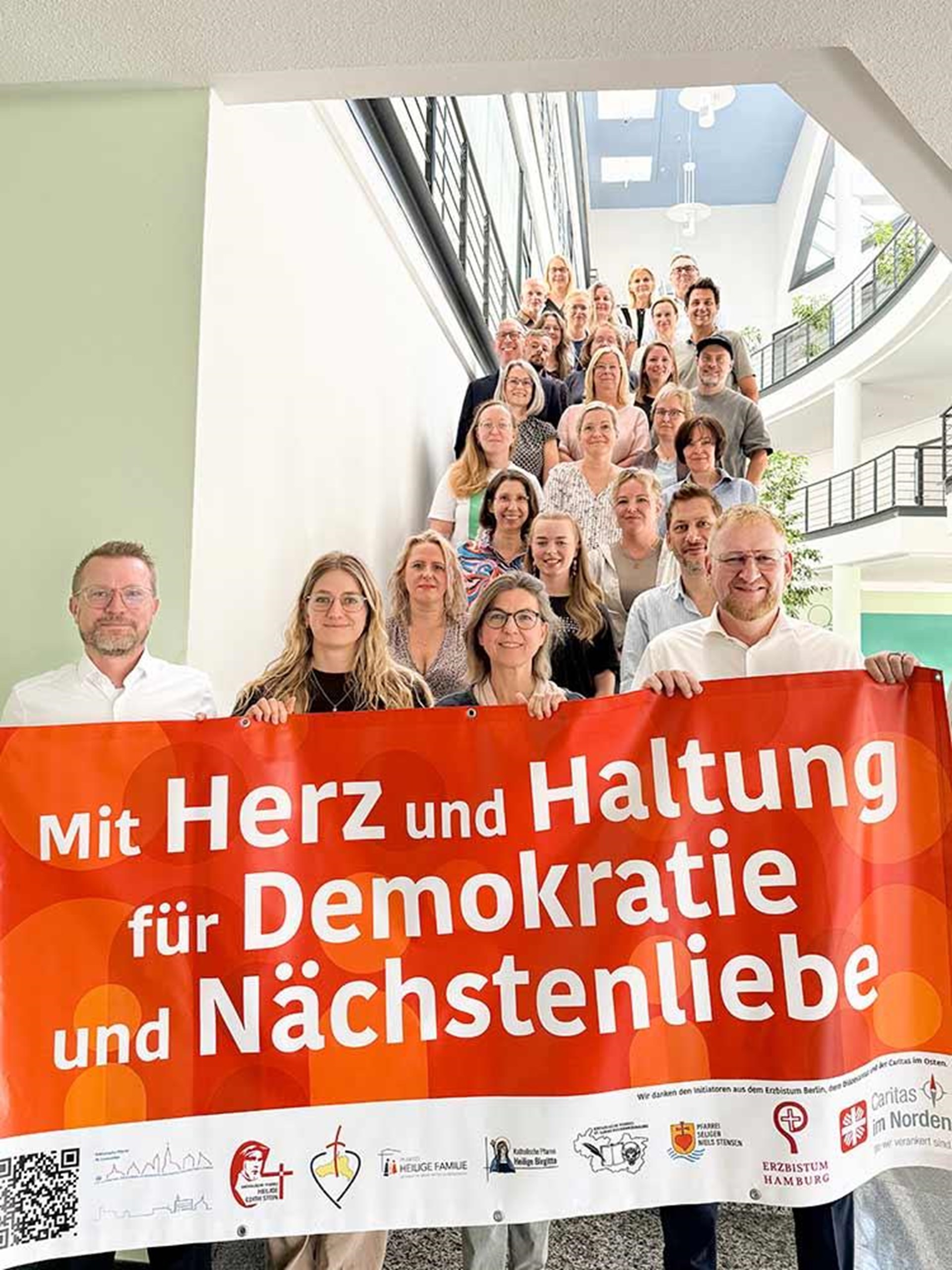 Herr Timmermann und Herr Meindl stehen gemeinsam mit den Mitarbeitern auf der Treppe und halten ein Schild mit der Aufschrift „Mit Herz und Haltung für Demokratie und Nächstenliebe” in den Händen.