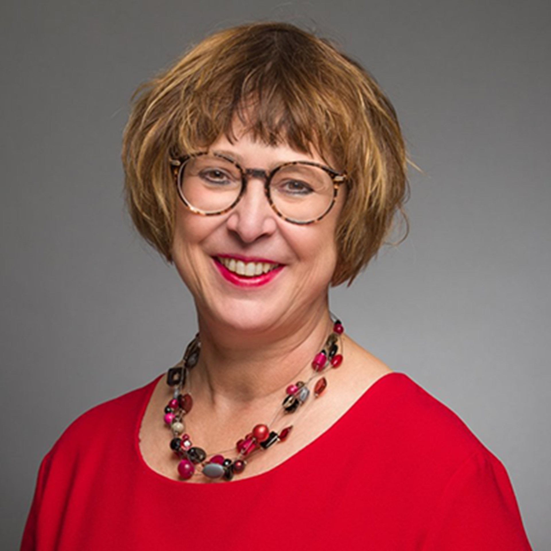 Lächelnde Frau mit braunen Haaren und Brille, die ein rotes T-Shirt und eine Halskette mit bunten Steinen trägt